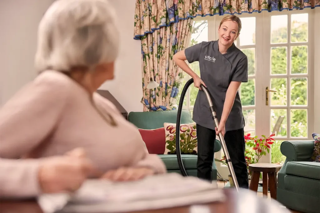 Carer cleaning a a living room, while an older lady is talking to her