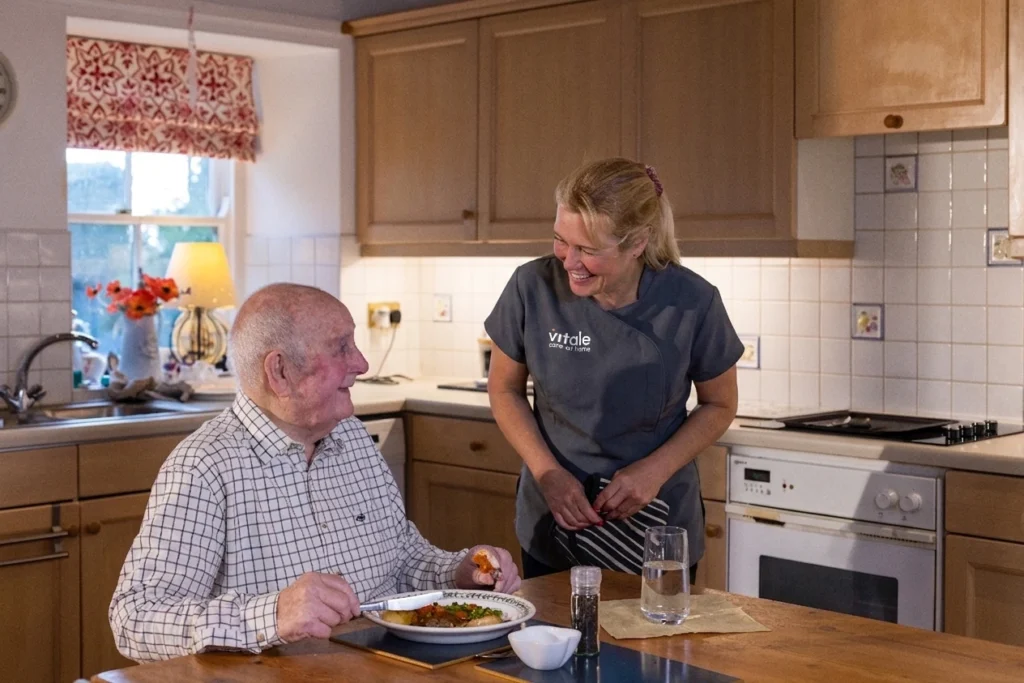 A Vitale Care Professional chatting to an elderly gentleman at his kitchen table enjoying a cup of tea